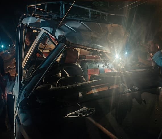 Illustration of a wrecked vehicle at night, lit by headlights, with bystanders gathered at the roadside.